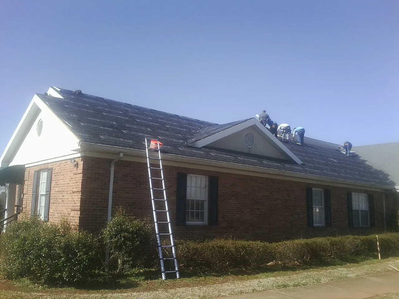 Crew tearing shingles off a brick ranch roof with a ladder set — Metal Roof Repair work by our crew in Uhrichsville