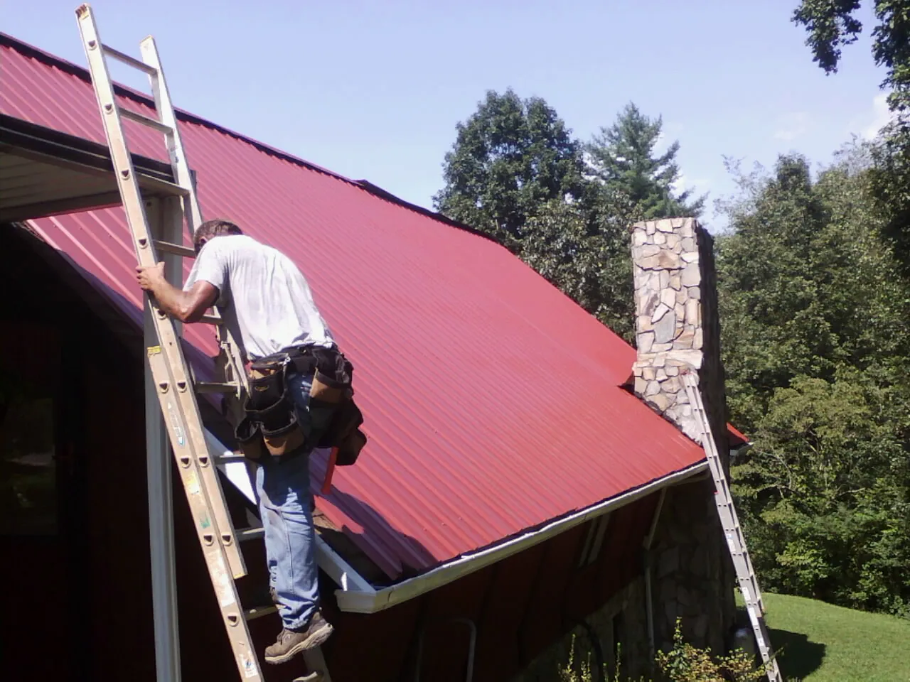 Roofer on a ladder beside a red metal roof and stone chimney — Soffit & Fascia Repair by Roofing Brothers in Uhrichsville